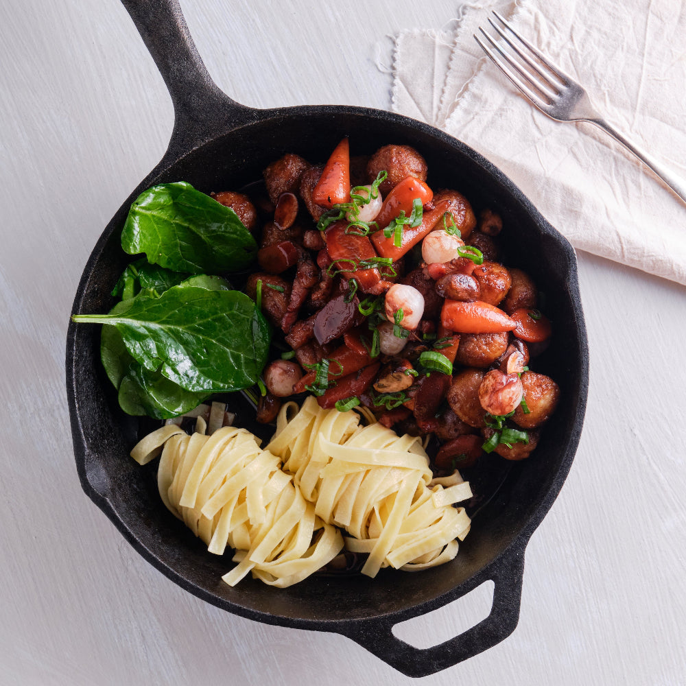 Black cast iron skillet with pasta, sautéed vegetables, and greens on a white surface.