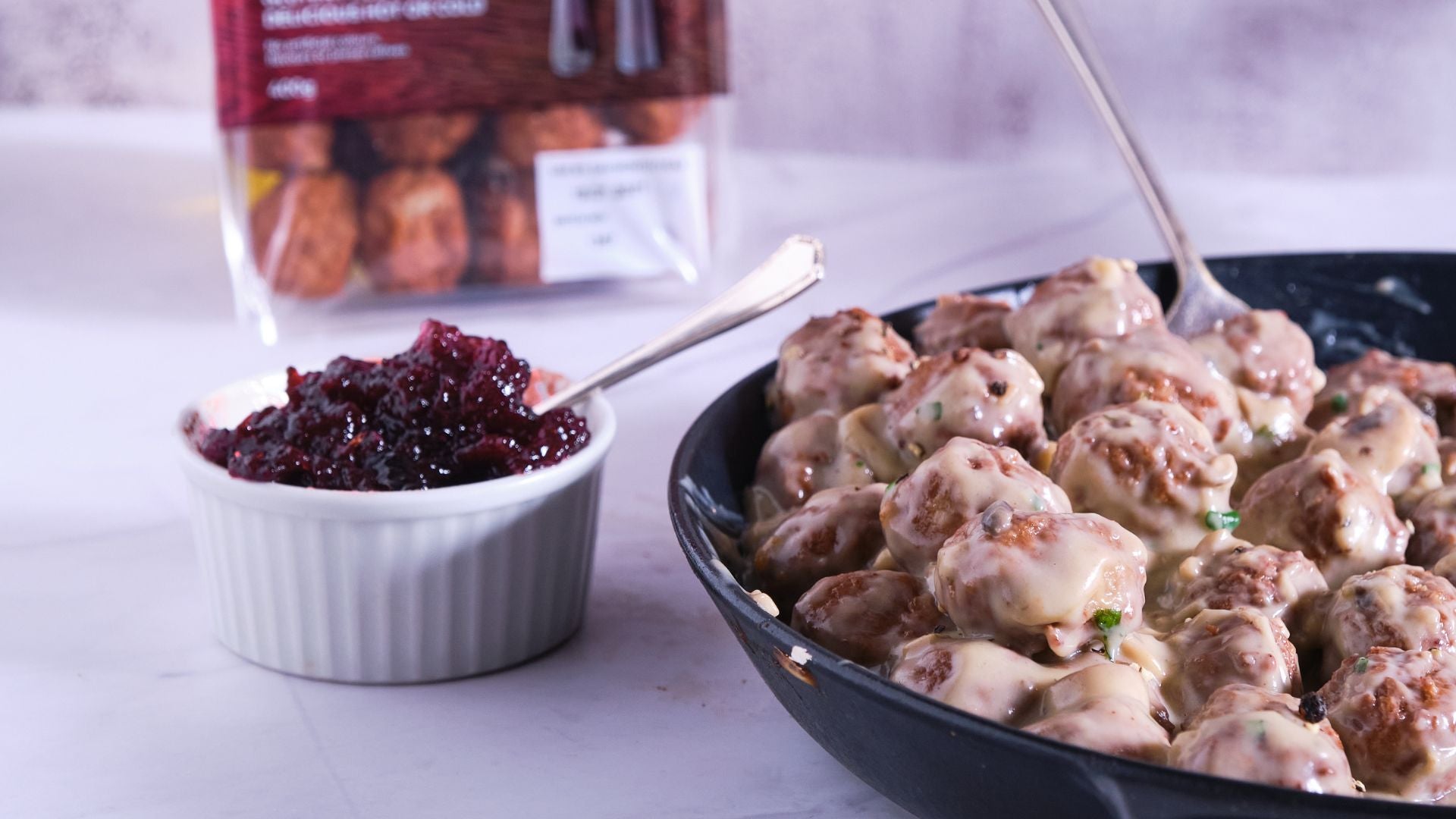 Skillet of meatballs with a side of lingonberry sauce on a white surface.