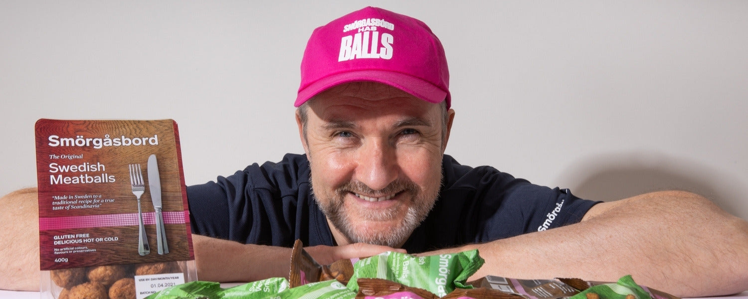 Man wearing a pink cap with 'Balls' printed on it, holding a package of Swedish meatballs.